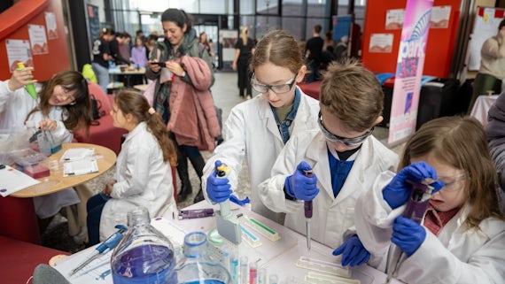 A group of children wearing lab coats transport liquid with pipettes.