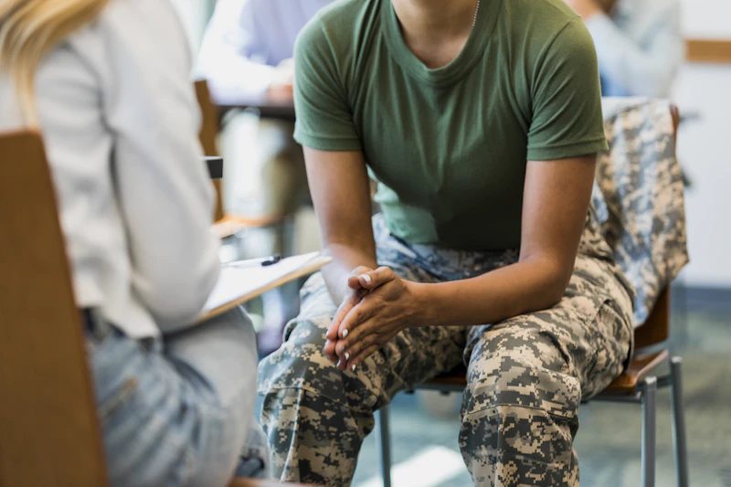 Person in army uniform, leaning forward talking to a medical professional holding a clipboard