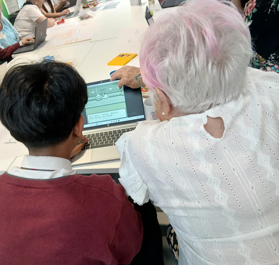 An older person and a teenager are facing away from the camera lens, looking at a laptop screen displaying pixel art. 